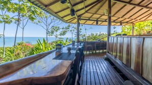 An outdoor dining area with wooden tables and chairs, a covered roof, and a view of the ocean.