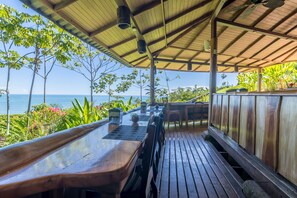 An outdoor dining area with wooden tables and chairs, a covered roof, and a view of the ocean.