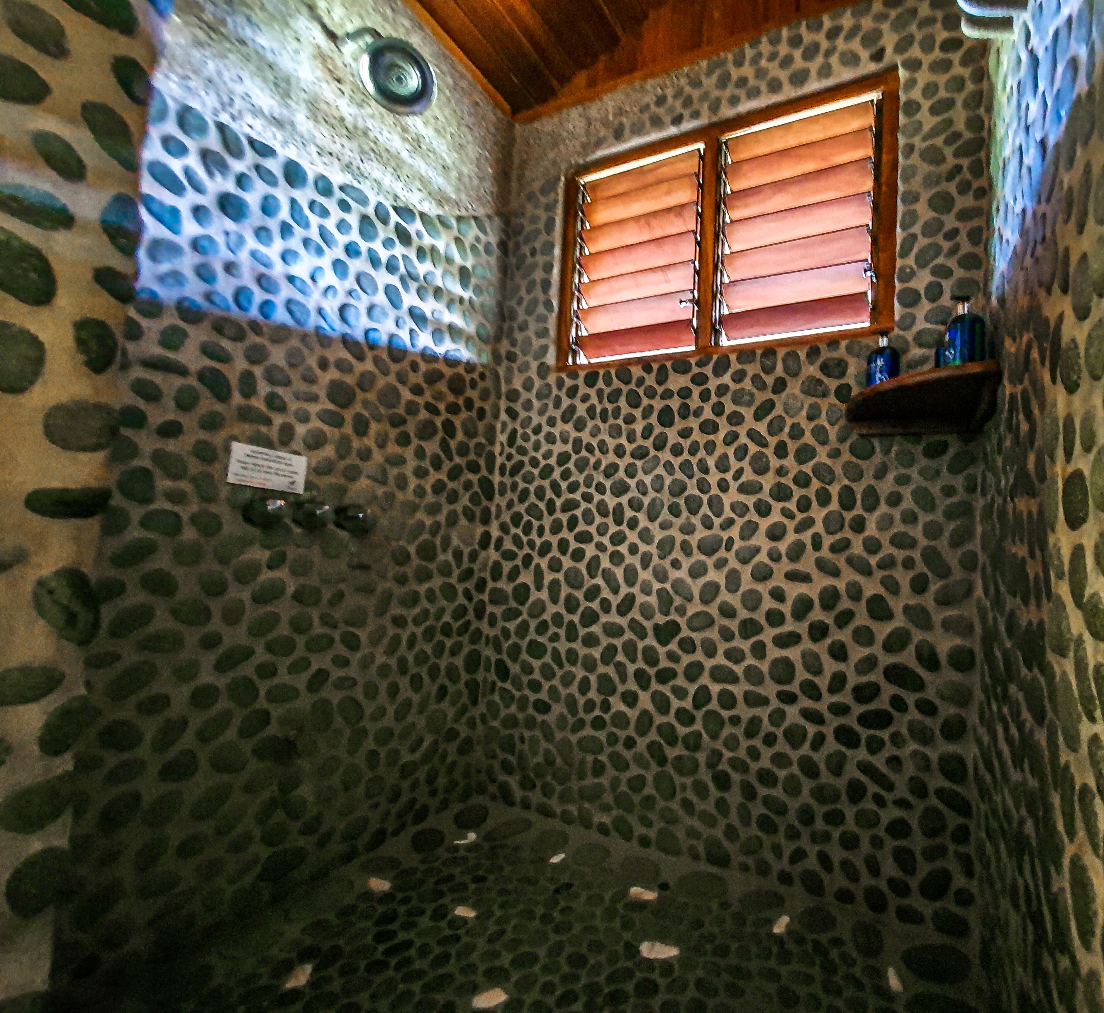 A stone-walled bathroom with a shower area, a window with blinds, and a shelf with bottles.