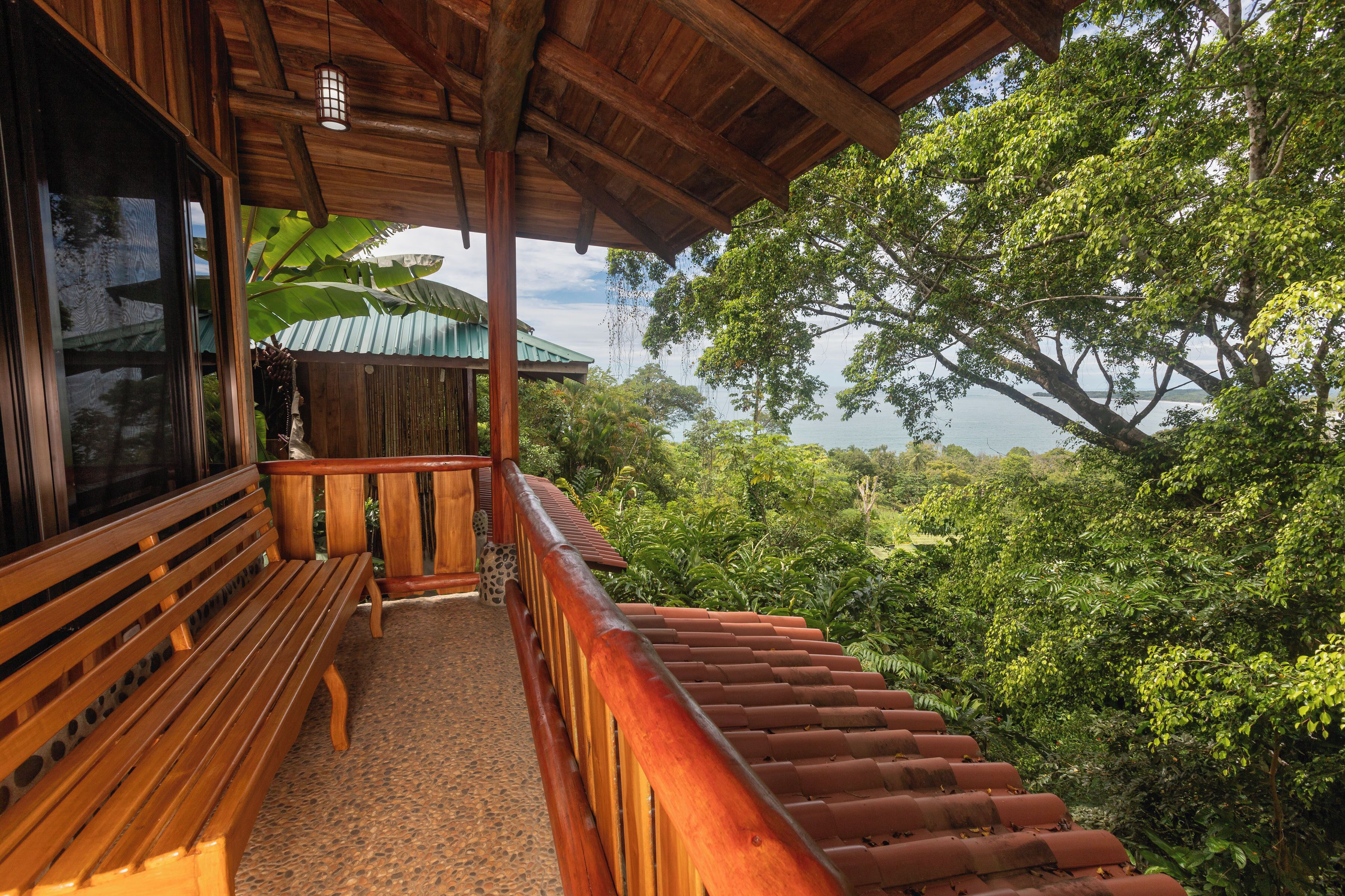 Un balcon en bois avec un banc, un escalier et une vue sur une végétation luxuriante et une étendue d’eau.