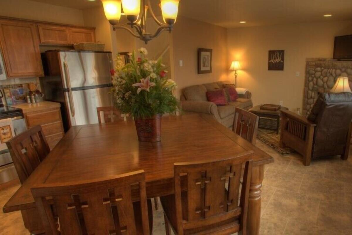 A kitchen with wooden cabinets, a stainless steel refrigerator, a dining table with chairs, and a flower vase.