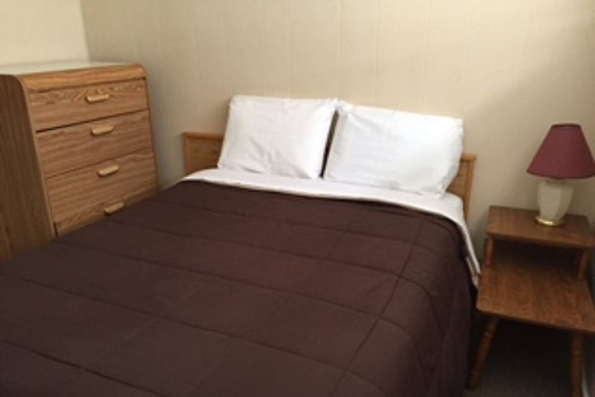 A single bed with a brown quilt, a wooden nightstand with a lamp, and a wooden dresser with drawers.