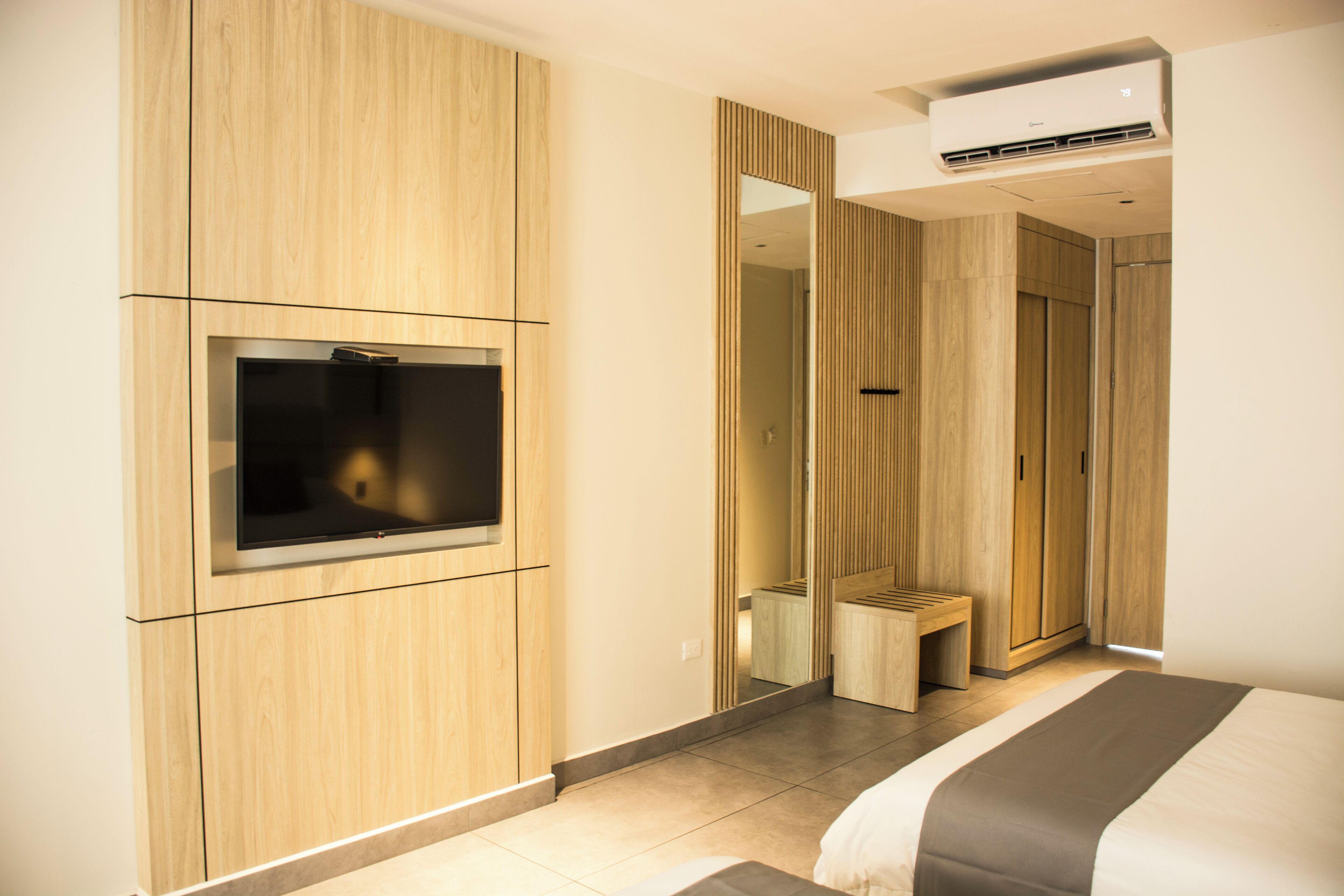 Habitación de hotel moderna con una televisión de pantalla plana montada en la pared, un pequeño banco y un espejo grande.