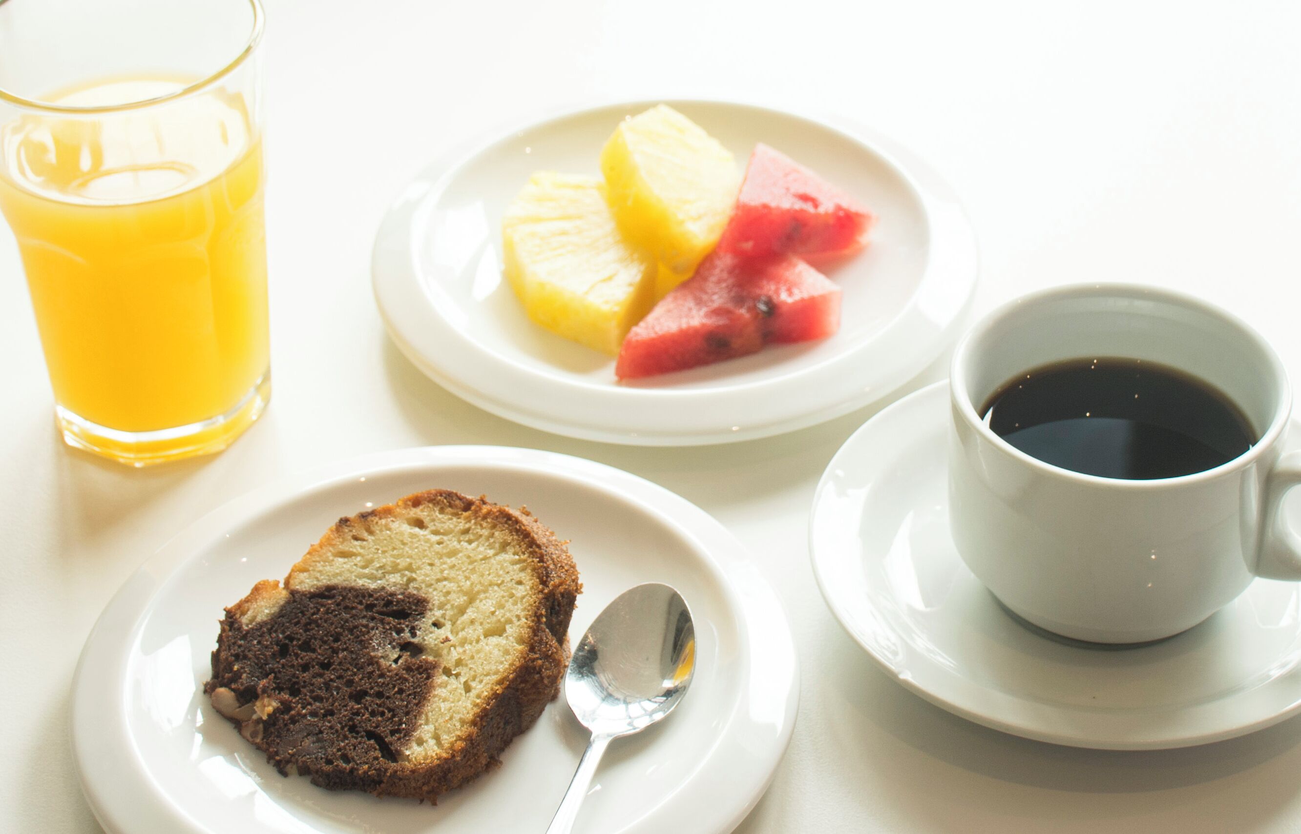 A breakfast setting with a slice of cake, a glass of orange juice, a plate with pineapple and watermelon, and a cup of coffee.