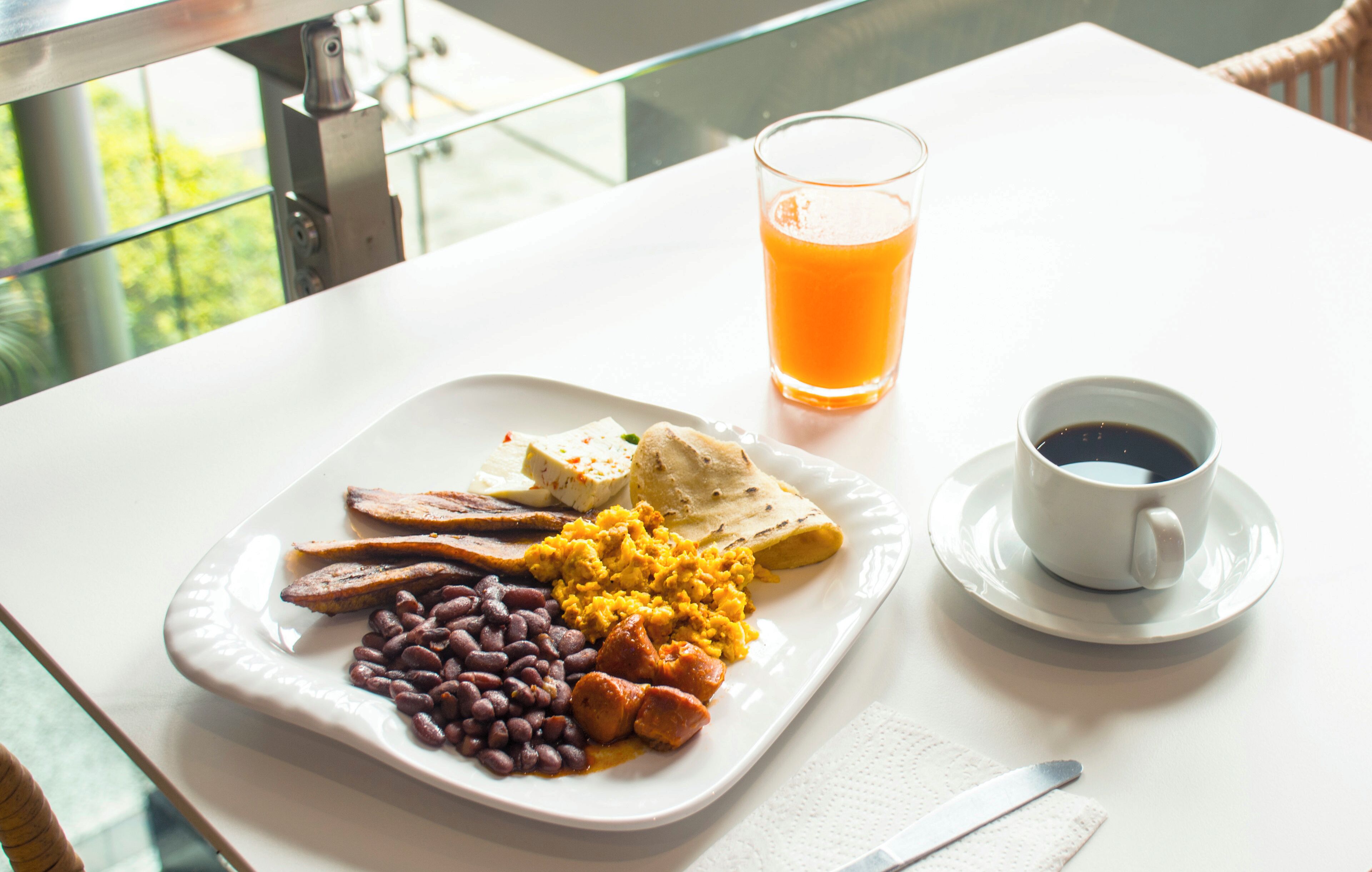 En tallerken med bønner, pølse og røræg, et glas appelsinjuice og en kop kaffe på et hvidt bord.
