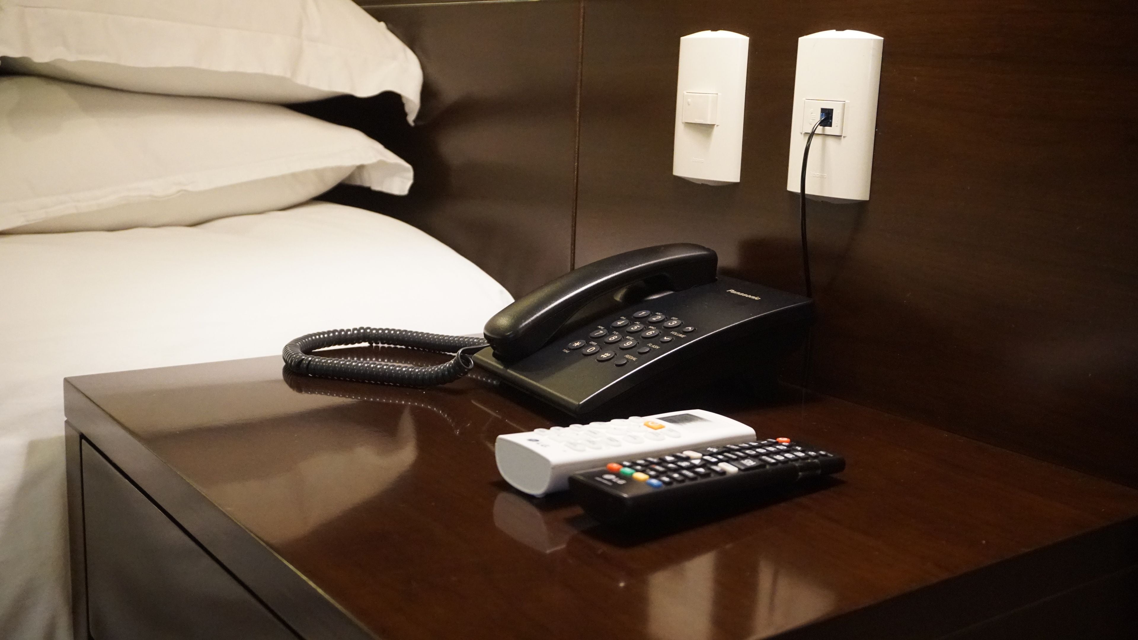 A hotel room with a bed, a bedside table with a telephone and a remote control.