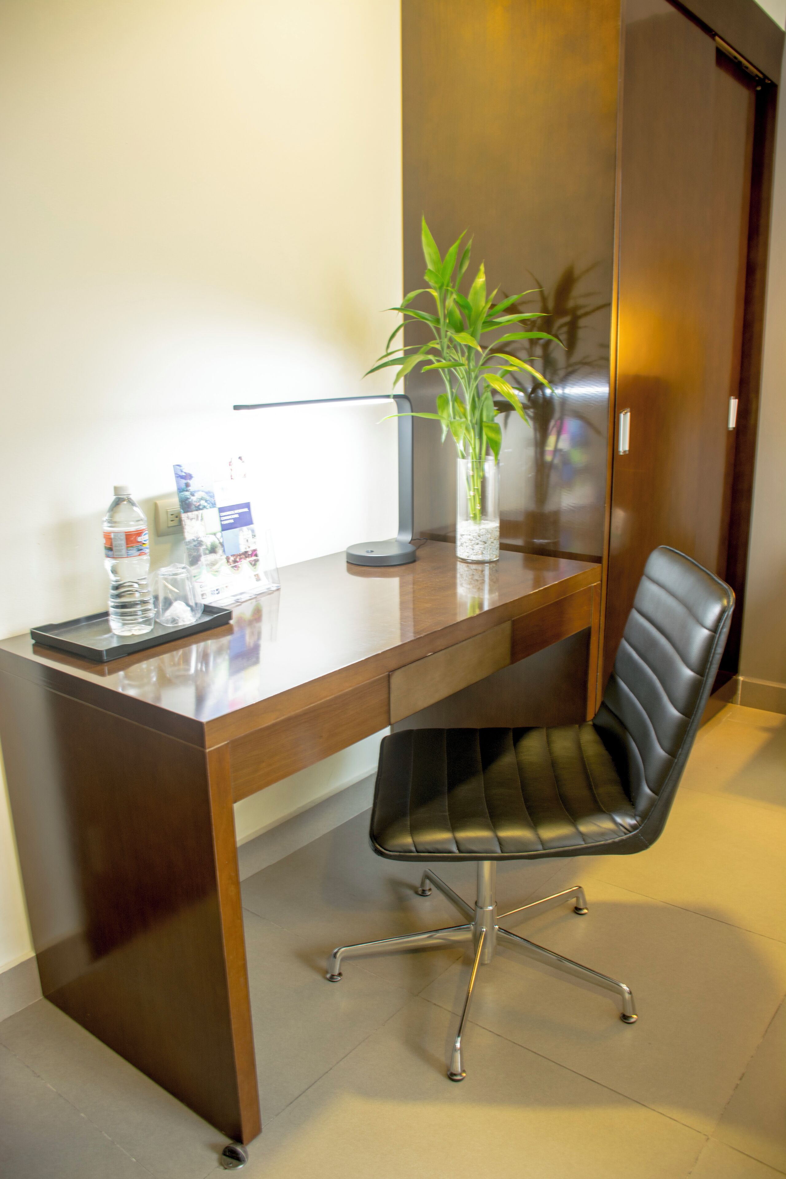 A wooden desk with a black office chair, a water bottle, a glass, and a potted plant.