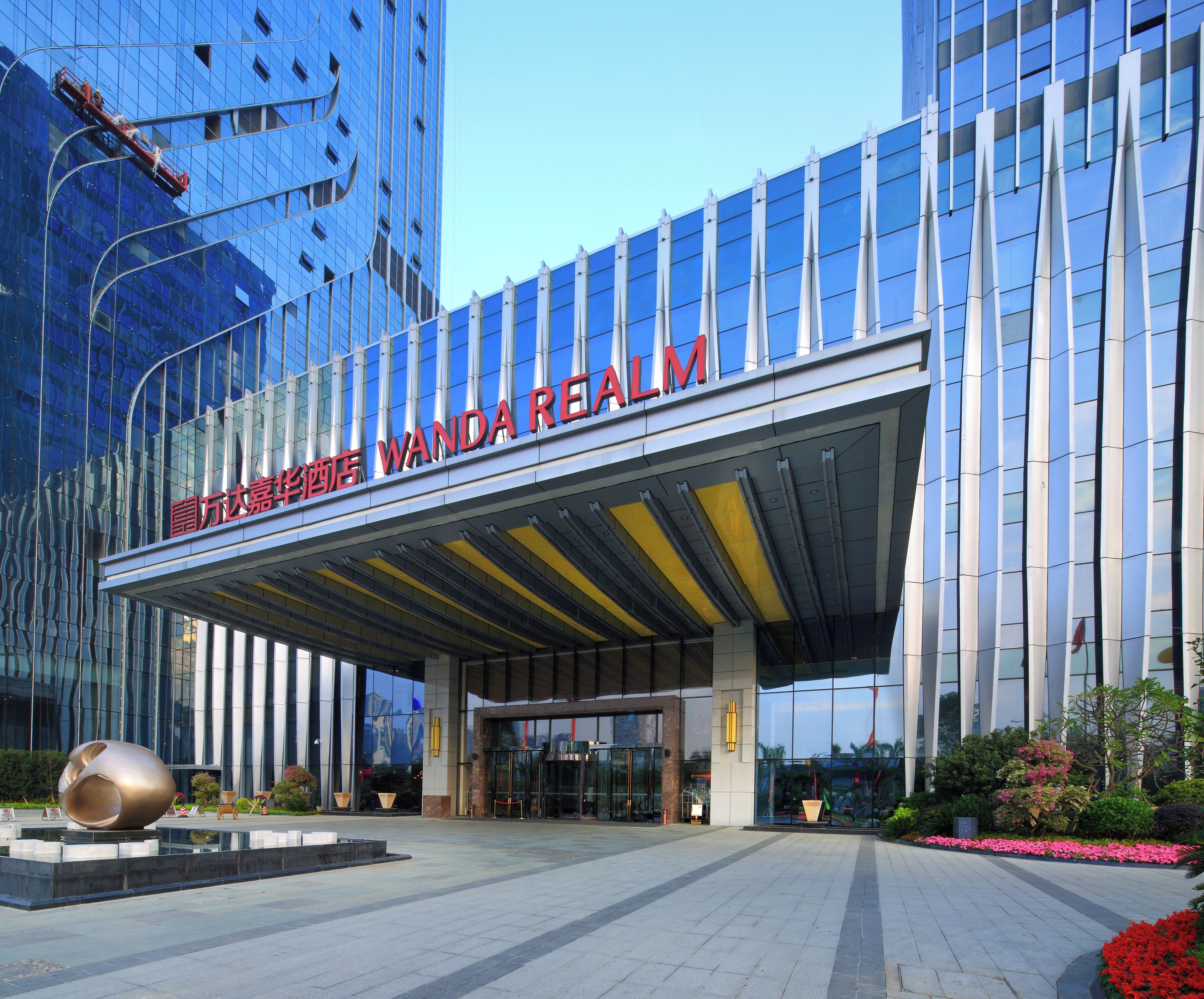 A modern hotel entrance with a clear sky, a large glass facade, and a prominent sign reading 'WANDA REALM'.