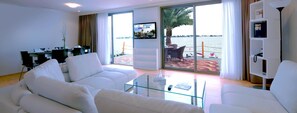 A modern living room with white sofas, a glass coffee table, and a TV screen displaying a resort.