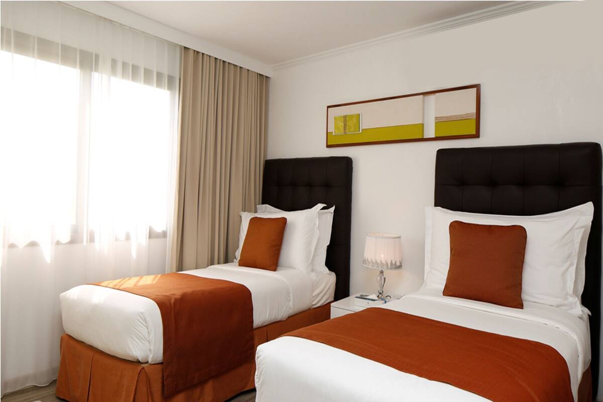 A hotel room with two beds, a window with sheer curtains, a bedside table with a lamp, and a framed artwork on the wall.