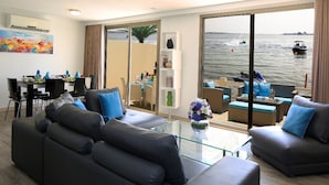 A modern living room with a glass coffee table, two sofas, and a dining area with chairs. A large window offers a view of the sea and boats. - Dana Beach Resort (Abqaiq)