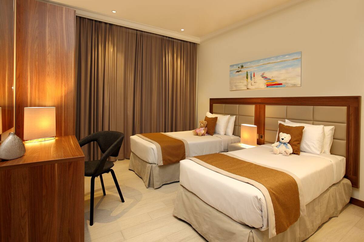 A hotel room with two beds, a desk, a chair, and a painting on the wall.