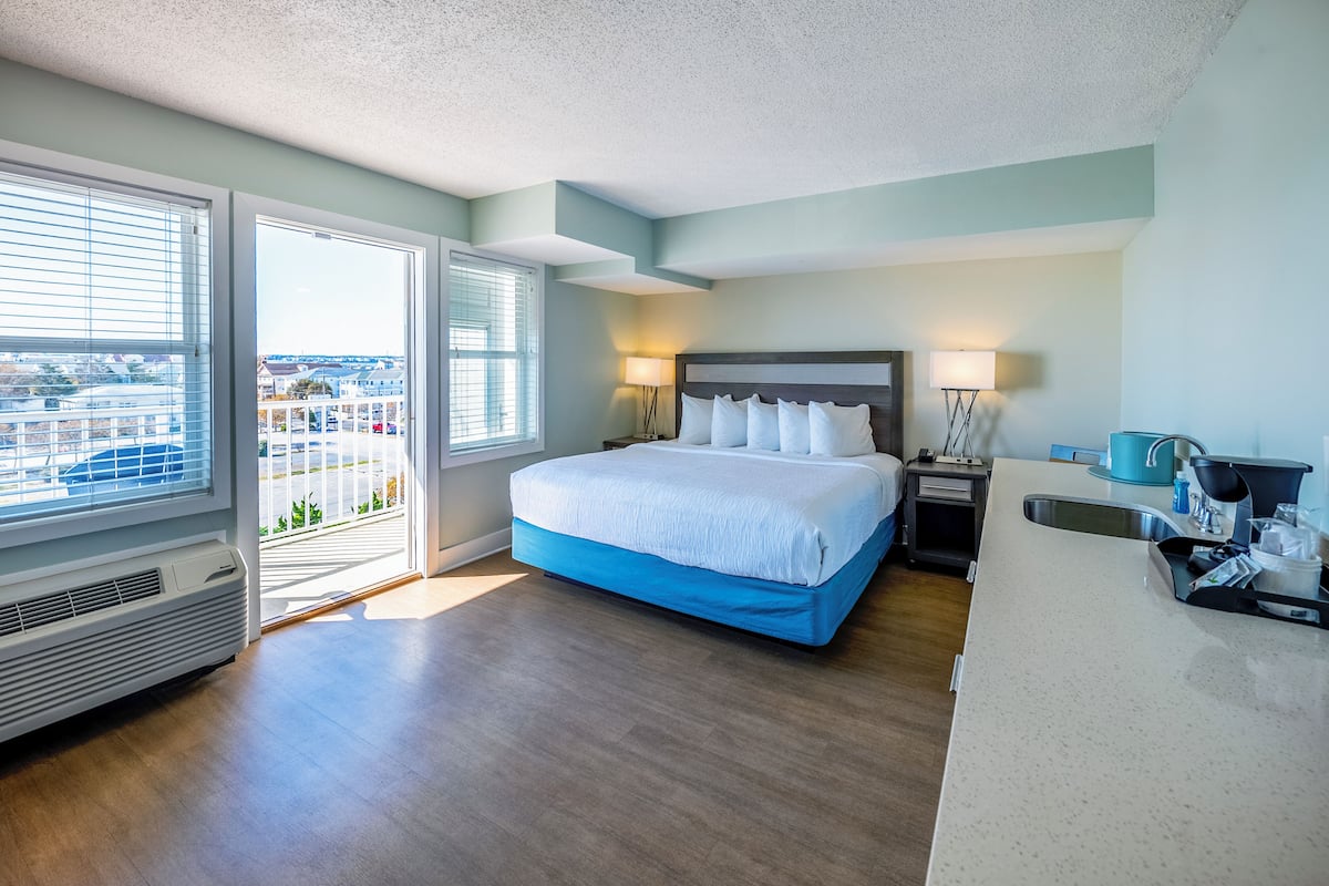 Habitación de hotel con cama, lámparas de noche, ventana con persianas, kitchenette y vistas al exterior.