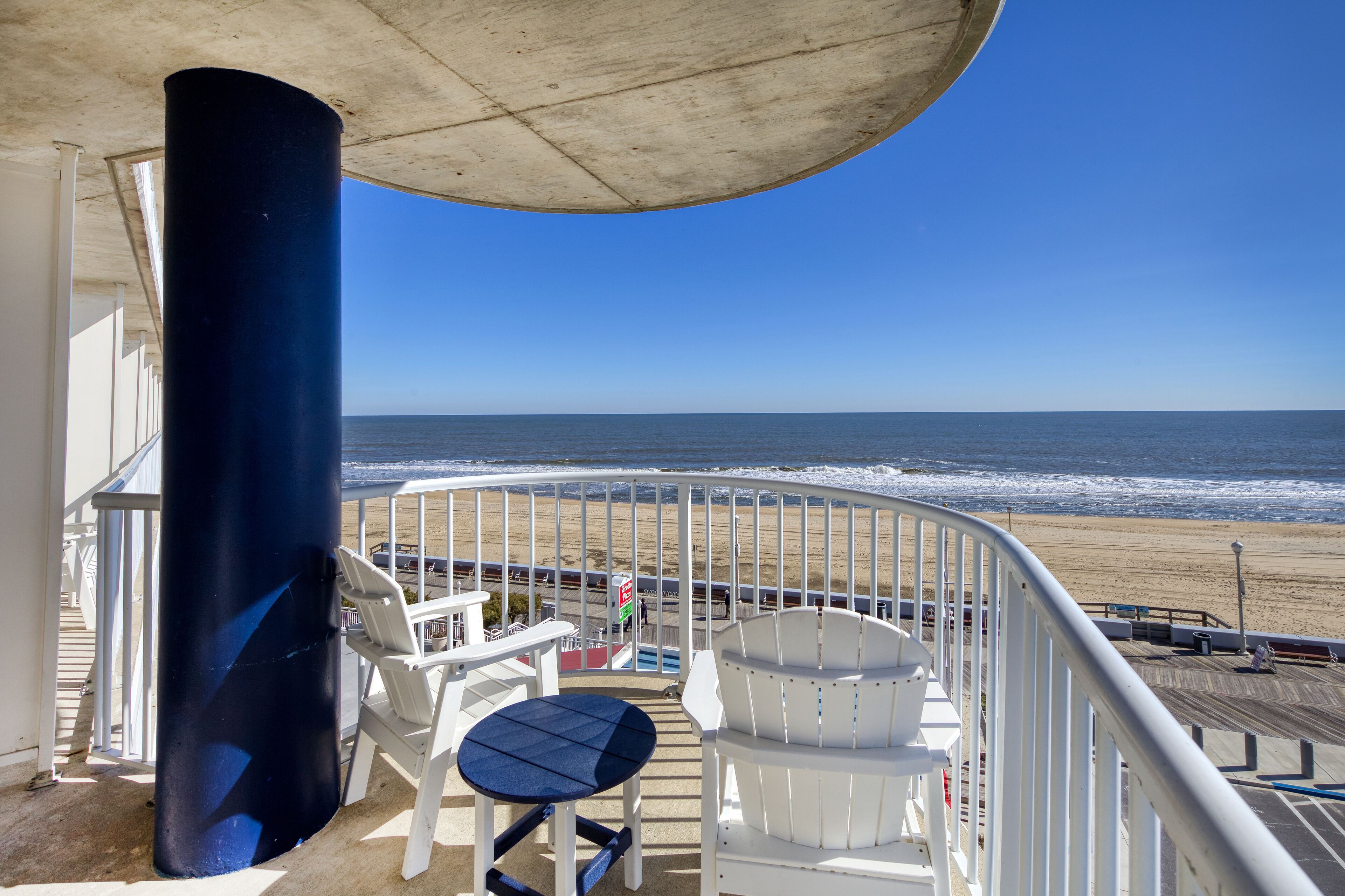 Ein Balkon mit Blick auf den Strand, weiße Stühle, ein Tisch und ein blauer Sessel.