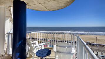 Ein Balkon mit Blick auf den Strand, weiße Stühle, ein Tisch und ein blauer Sessel.