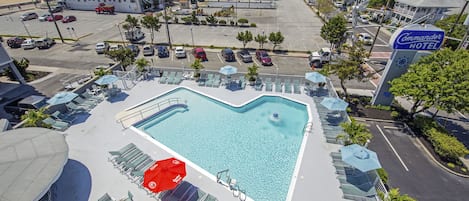 Et hotel med en stor swimmingpool omgivet af liggestole og parasoller, en parkeringsplads samt udsigt til stranden og havet.