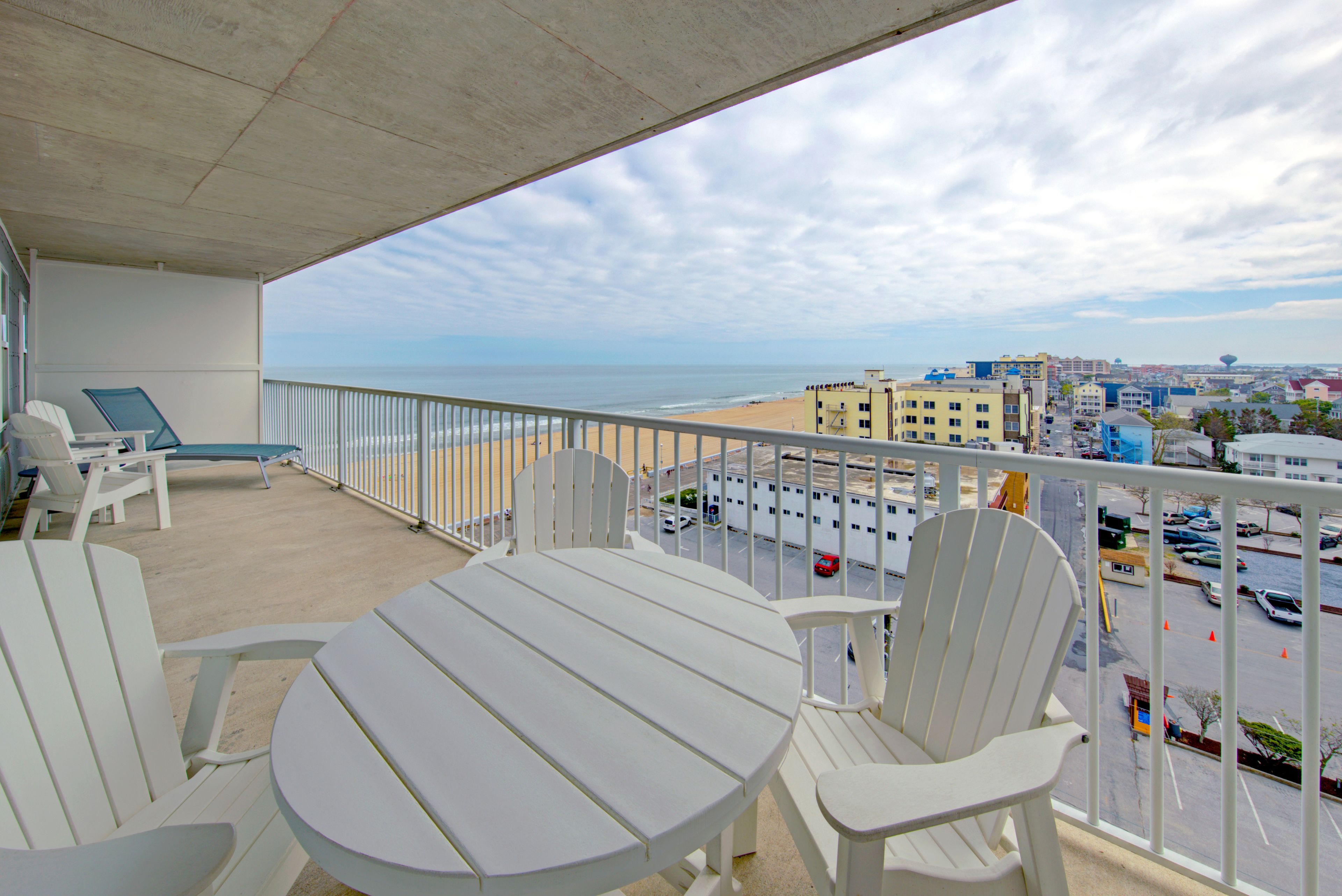 A balcony with a table and chairs, overlooking a beach and a parking lot.