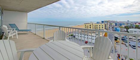 A balcony with a table and chairs, overlooking a beach and a parking lot.