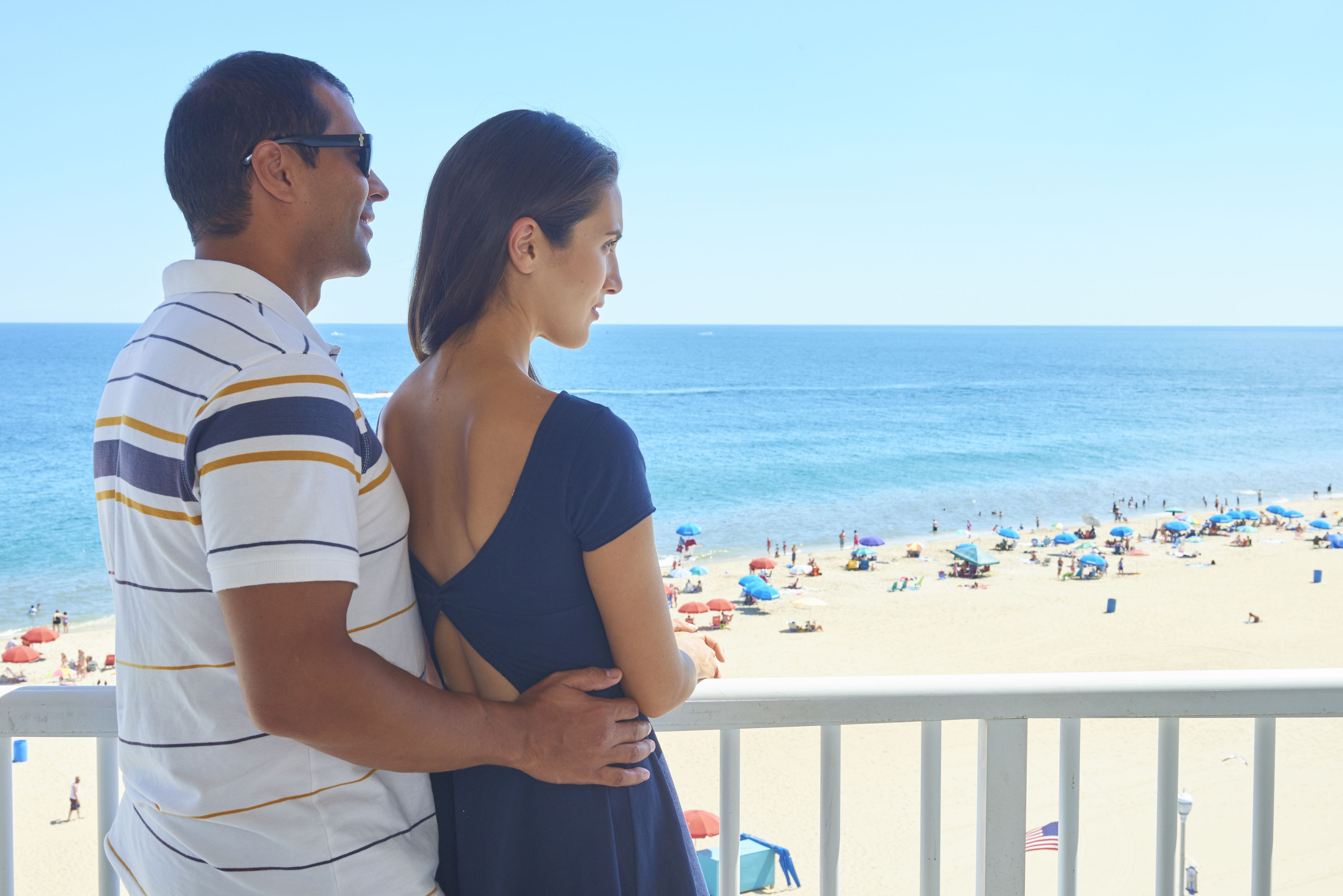 Una coppia in piedi su un balcone con vista su una spiaggia, ombrelloni e persone.