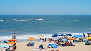 Une plage où des gens se détendent sous des parasols, avec une chaise de maître-nageur et un bateau sur l’eau.