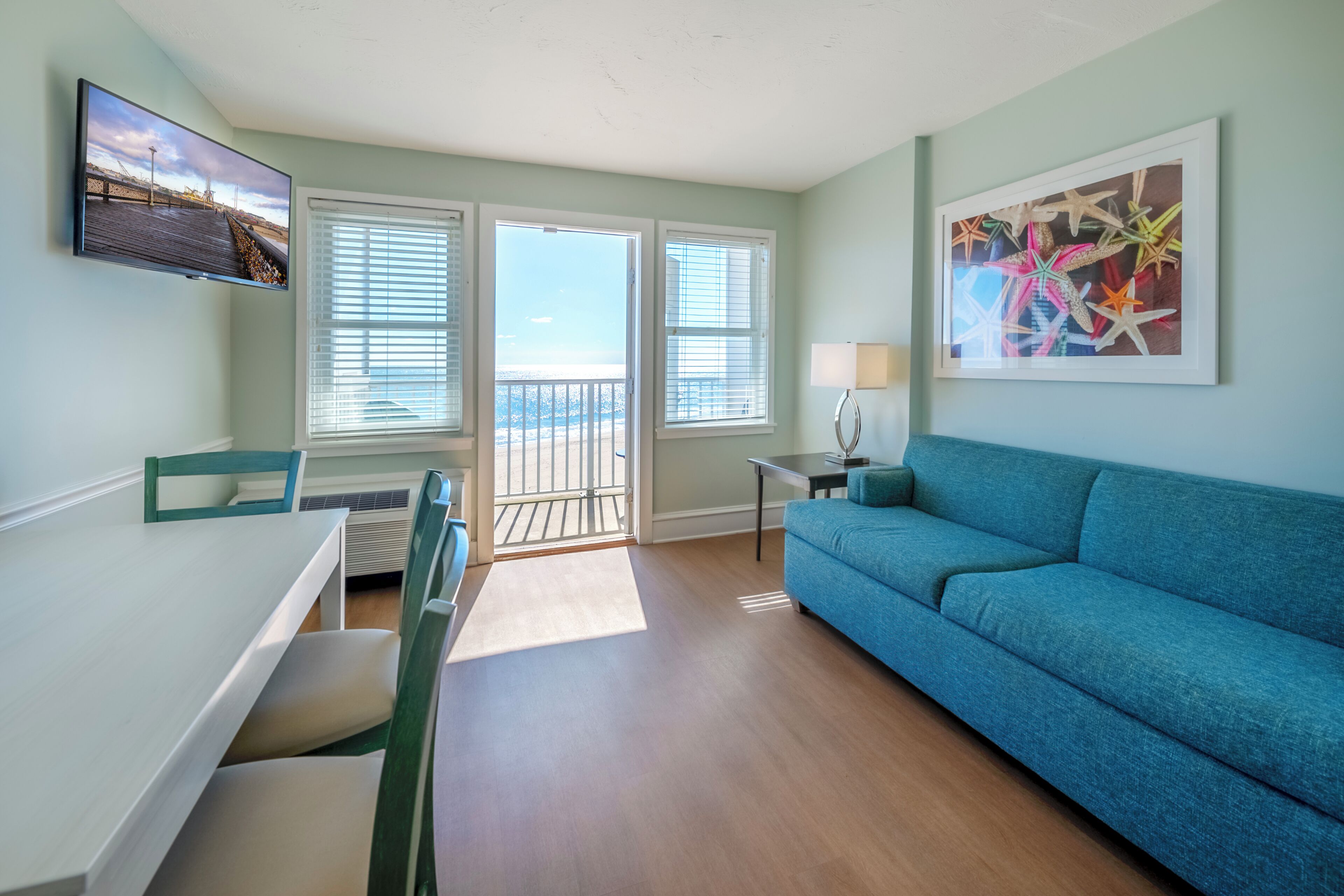 A living room with a blue sofa, a dining table with chairs, a TV mounted on the wall, and a balcony with a view.