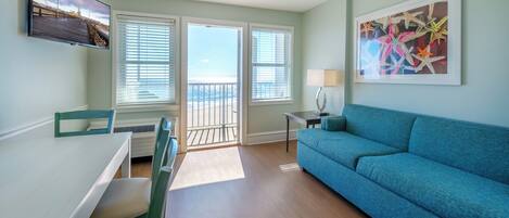 A living room with a blue sofa, a dining table with chairs, a TV mounted on the wall, and a balcony with a view.