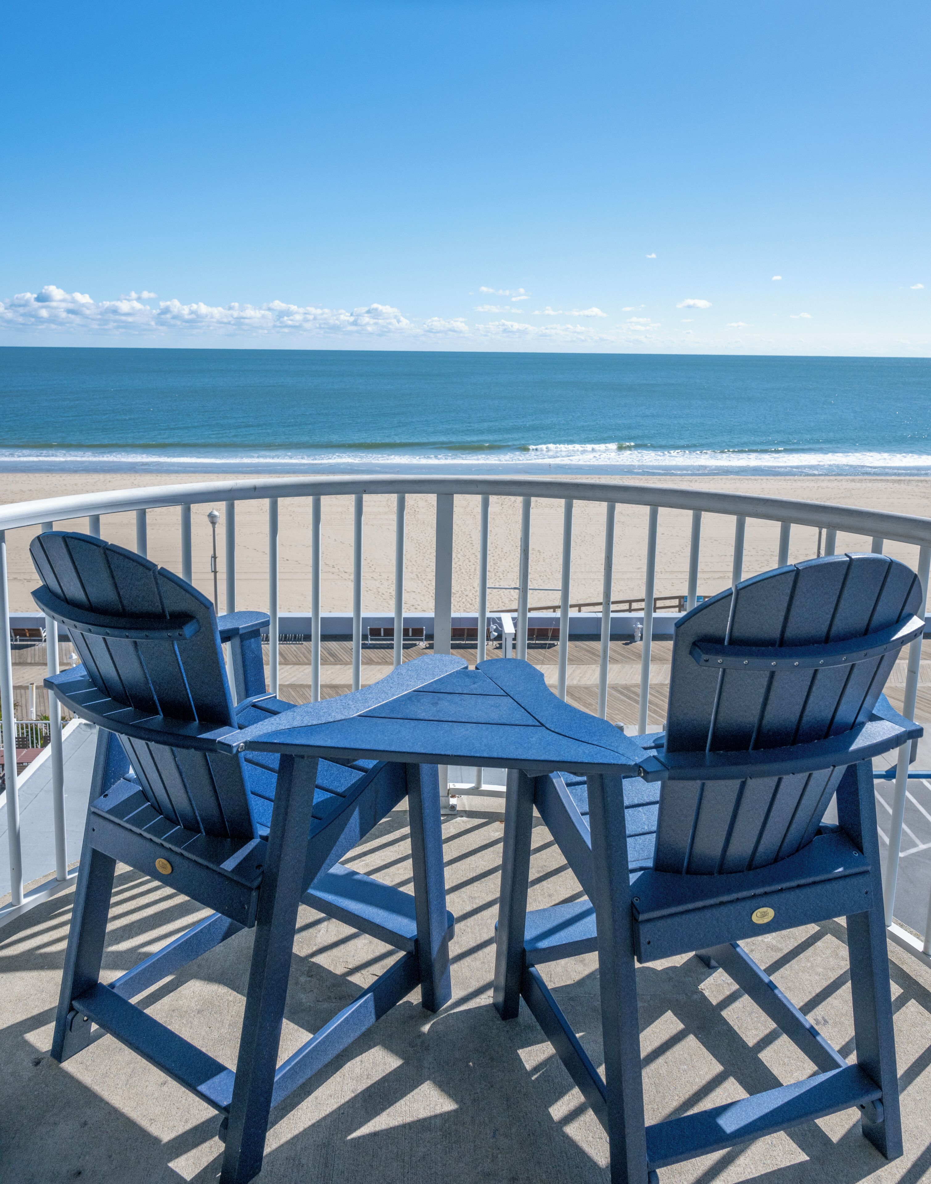 A balcony with two blue Adirondack chairs and a small table, overlooking a beach with waves.