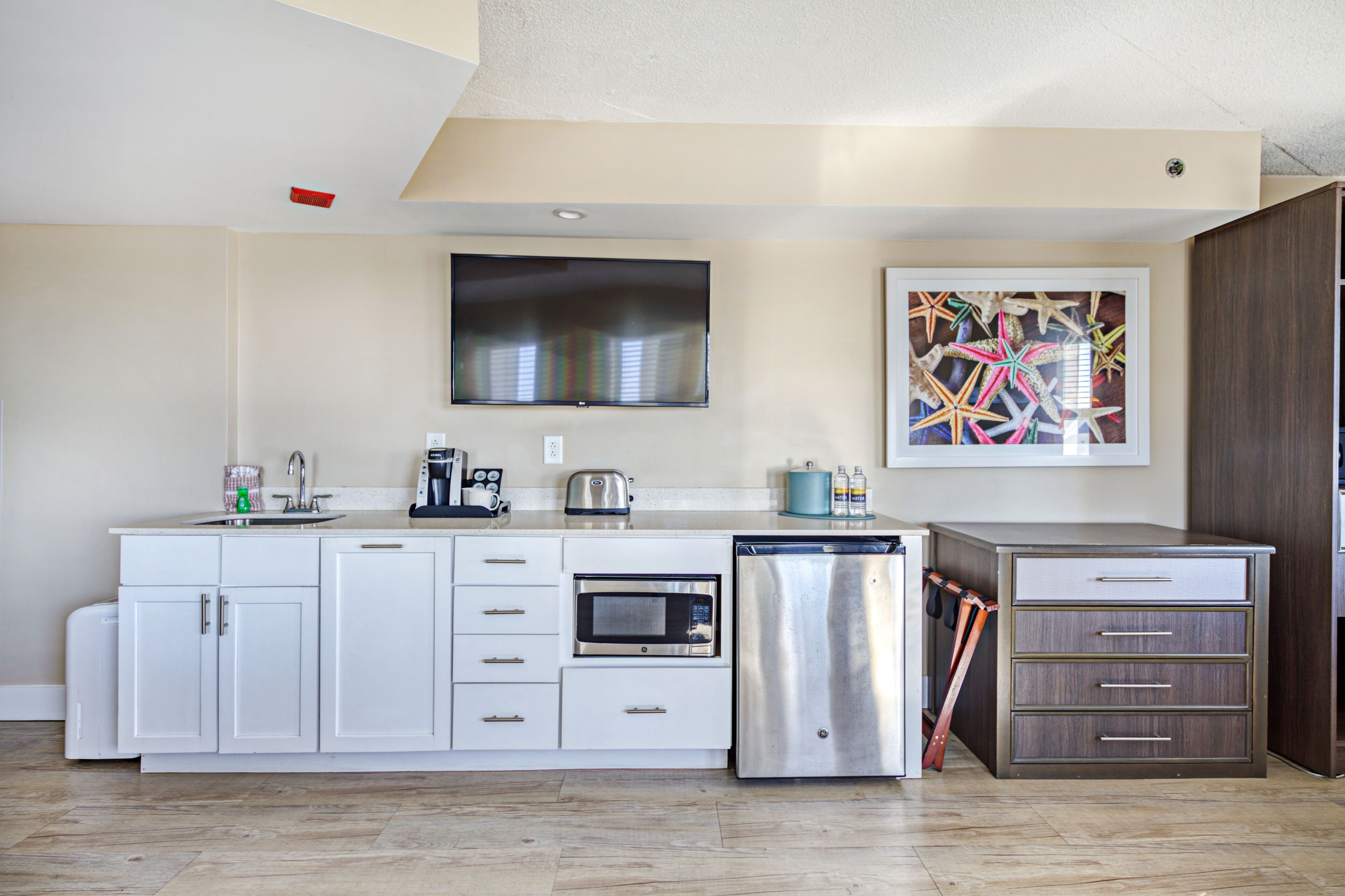 A modern kitchen with white cabinetry, a built-in microwave, a stainless steel refrigerator, and a flat-screen TV mounted above the counter.