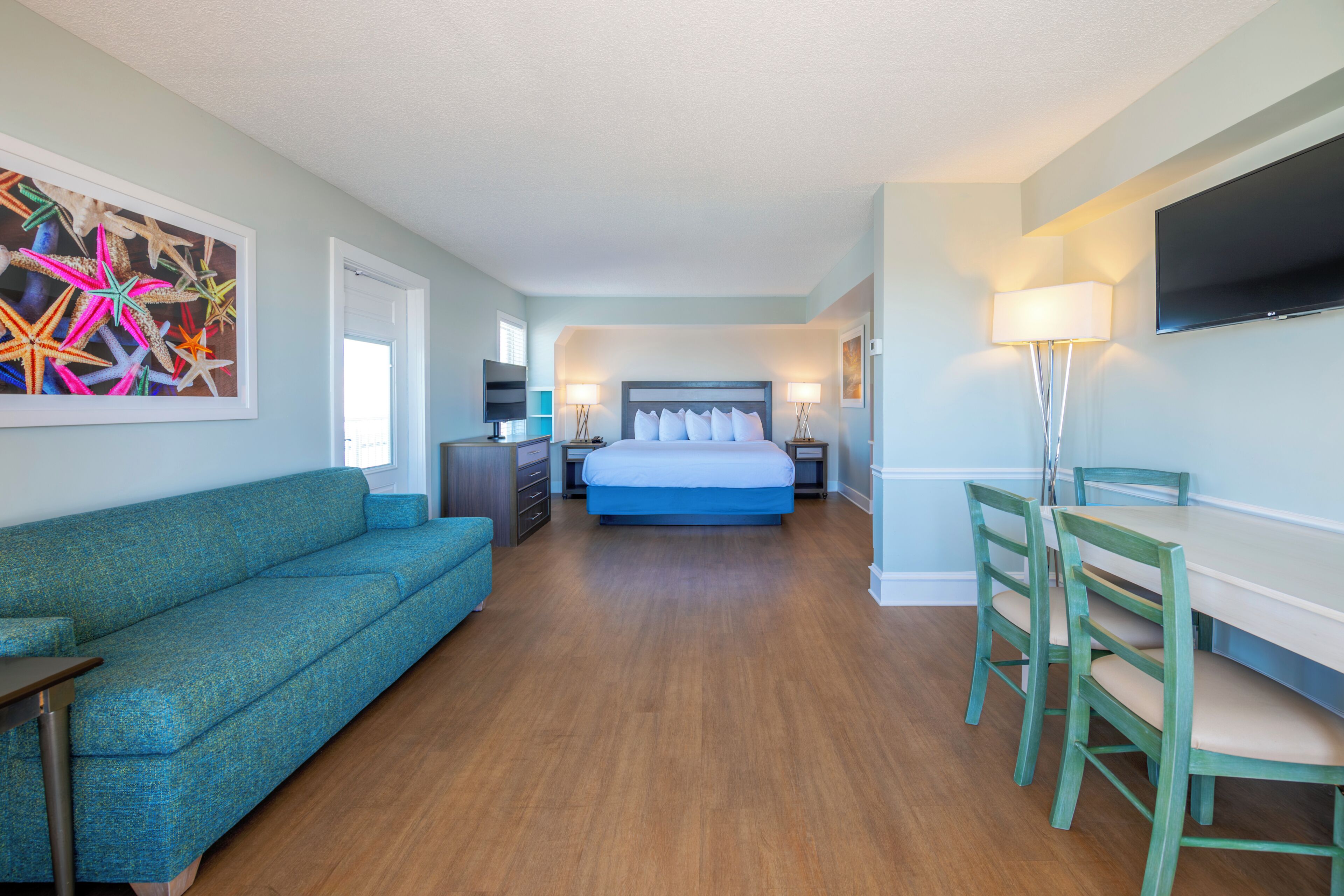 A hotel room with a bed, a sofa, a dining table with chairs, a TV, and artwork on the wall.