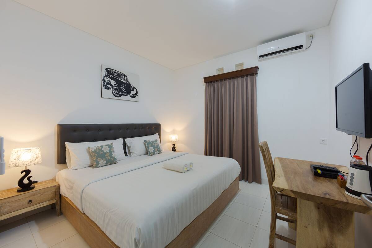 Ein Hotelzimmer mit Bett, Nachttisch, Lampe, Klimaanlage und Schreibtisch mit Stuhl.