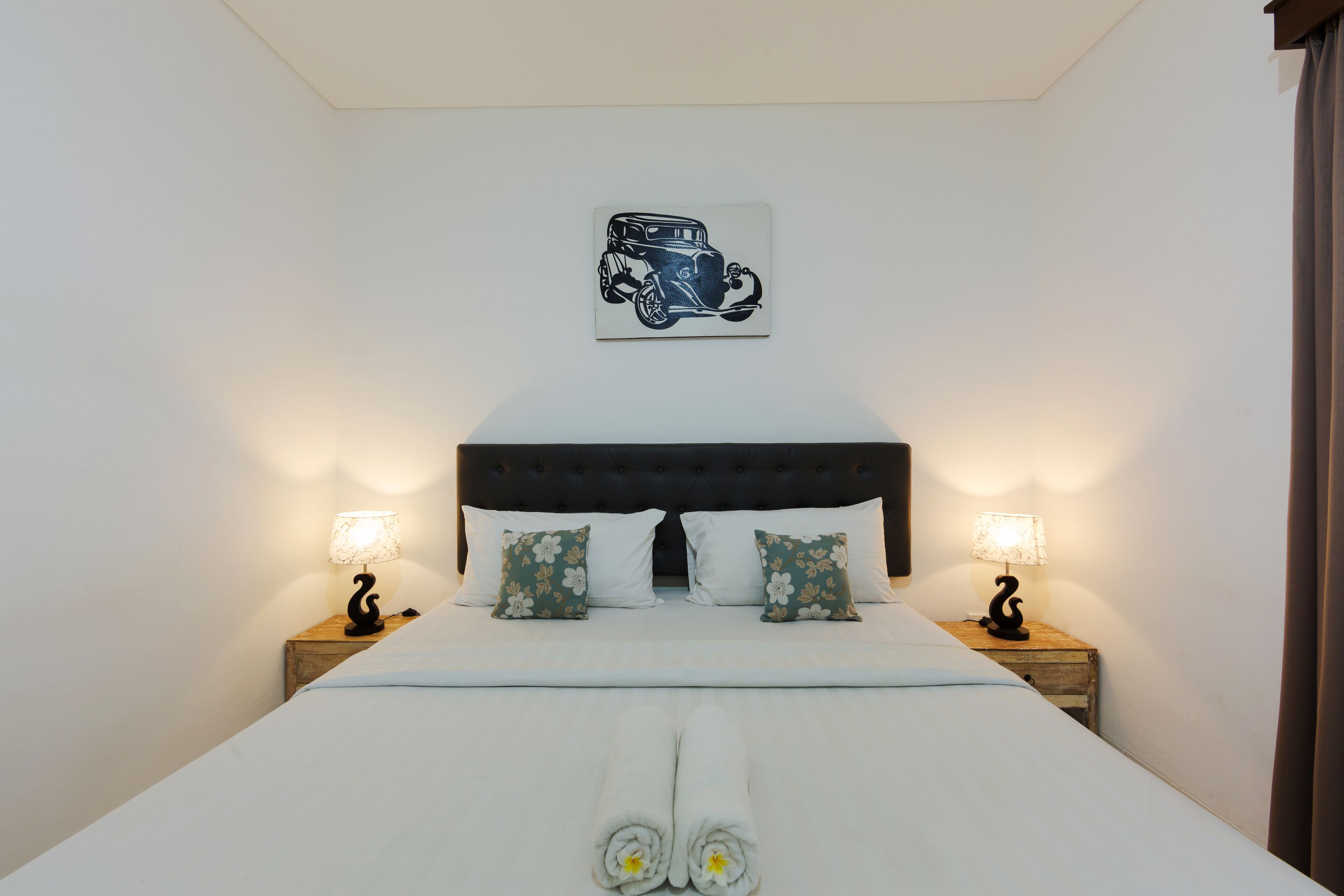 Ein ordentlich bezogenes Bett mit weißen Leinen und zwei Kissen, ein Wandbild mit einem Oldtimer sowie je eine Nachttischlampe auf Holzbeistelltischen.