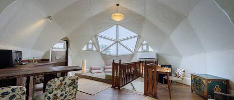 Um loft espaçoso com pé-direito alto, uma mesa de jantar de madeira, um sofá e uma escada que dá acesso a um andar superior.