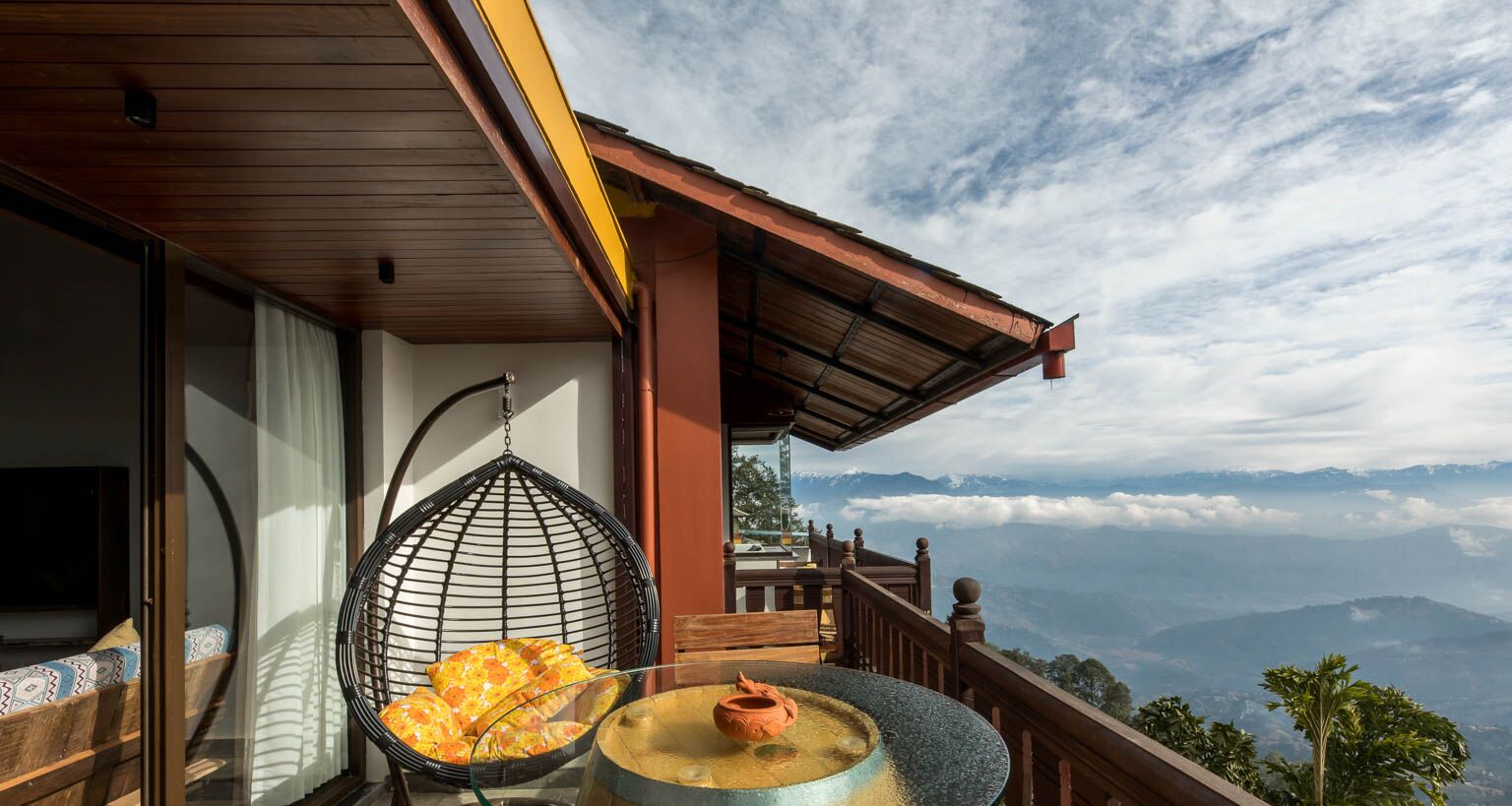 A balcony with a hanging chair, a table with a teapot, and a view of distant mountains.
