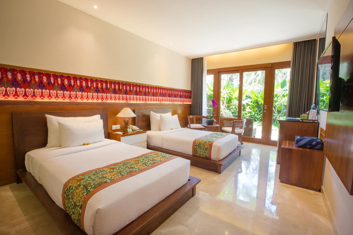 Quarto de hotel com duas camas, uma escrivaninha, uma cadeira e uma janela grande com vista para o verde.