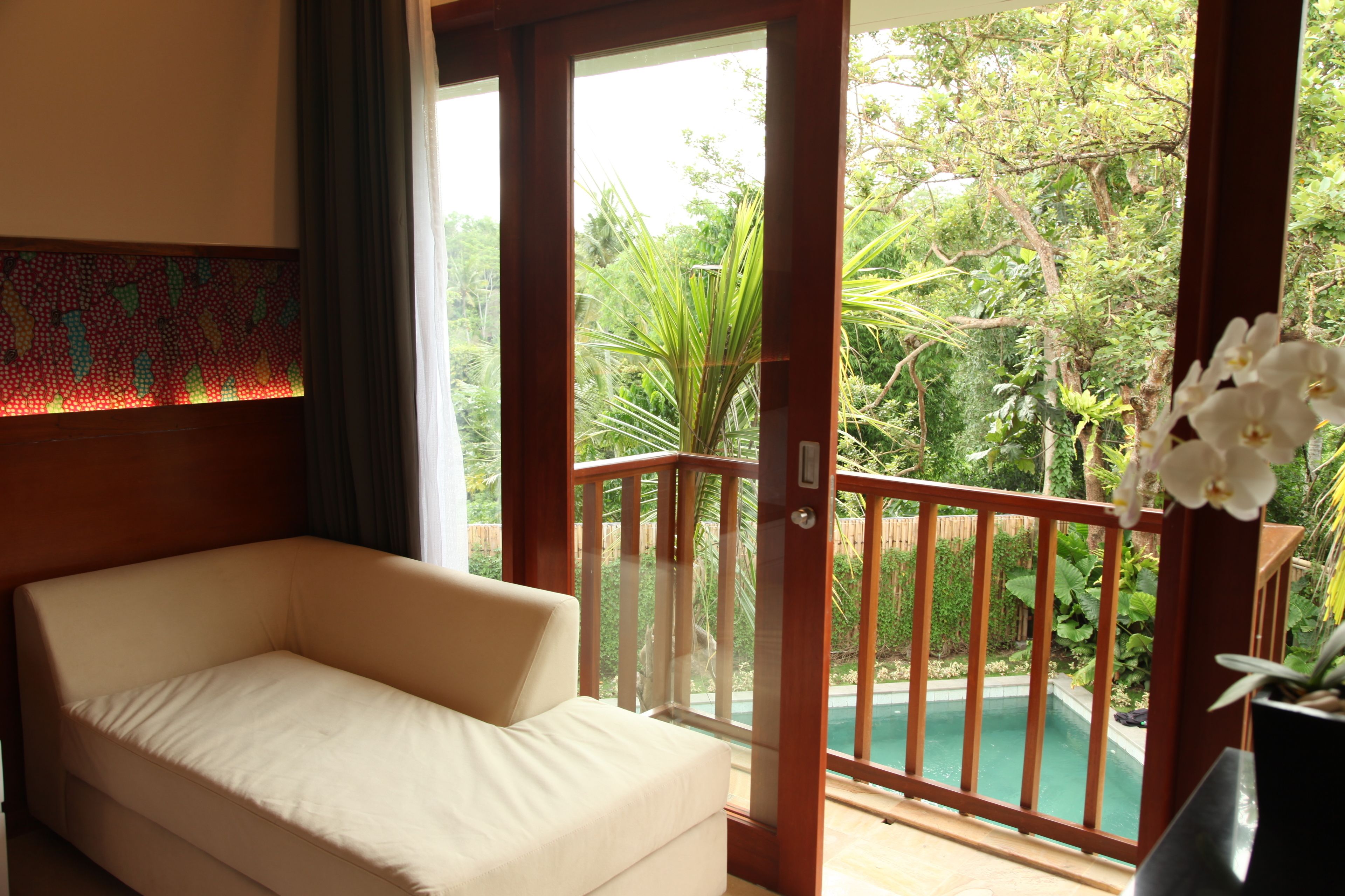 A room with a beige sofa, a wooden door, and a view of a pool and greenery.