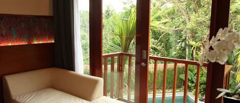 A room with a beige sofa, a wooden door, and a view of a pool and greenery.
