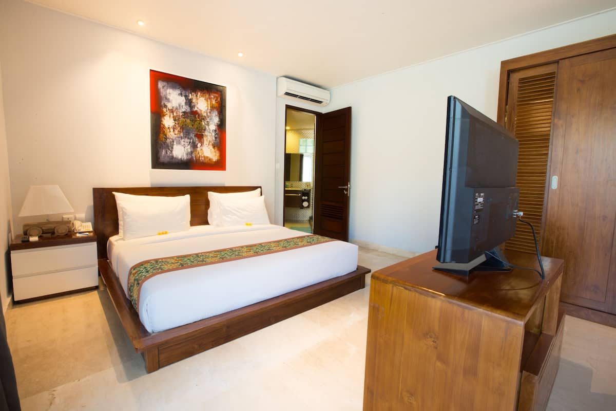 Quarto de hotel com uma cama, televisão sobre um móvel de madeira, mesa de cabeceira com abajur e um aparelho de ar condicionado na parede.