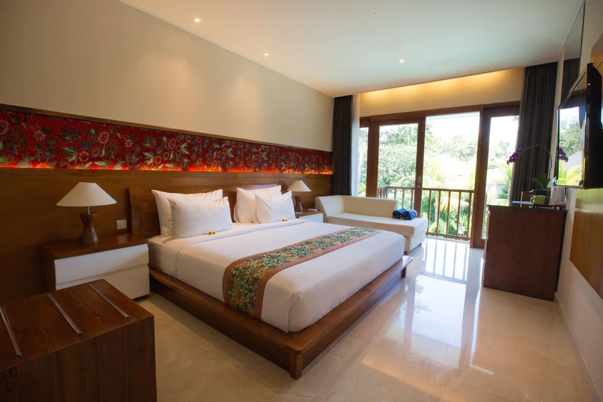 Um quarto espaçoso com uma cama grande, cabeceira de madeira, mesa de cabeceira, escrivaninha e varanda com vista para o verde.