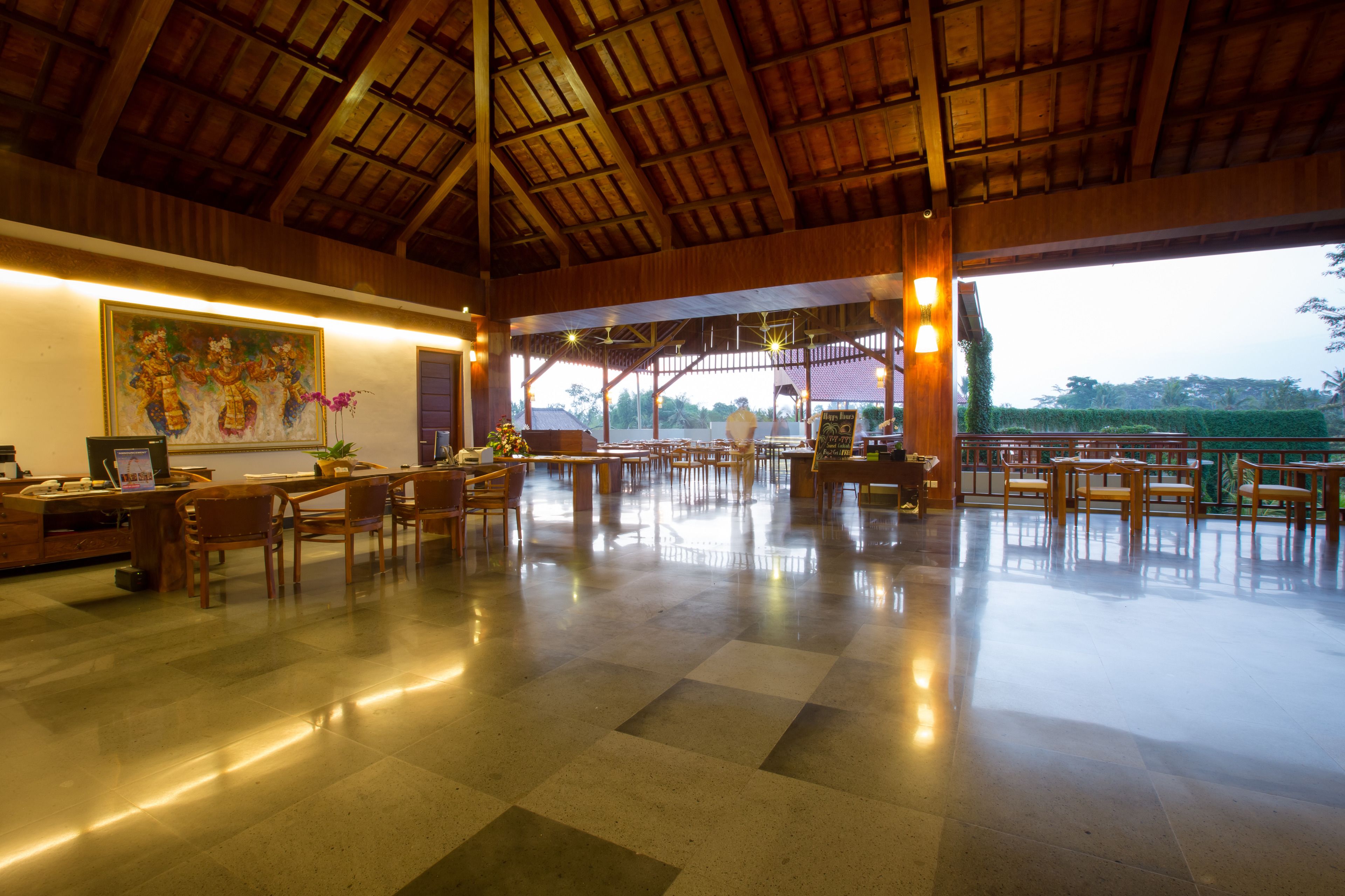 A spacious indoor area with a high ceiling and wooden beams, featuring a large painting on the wall, multiple tables and chairs, and a view of greenery outside.