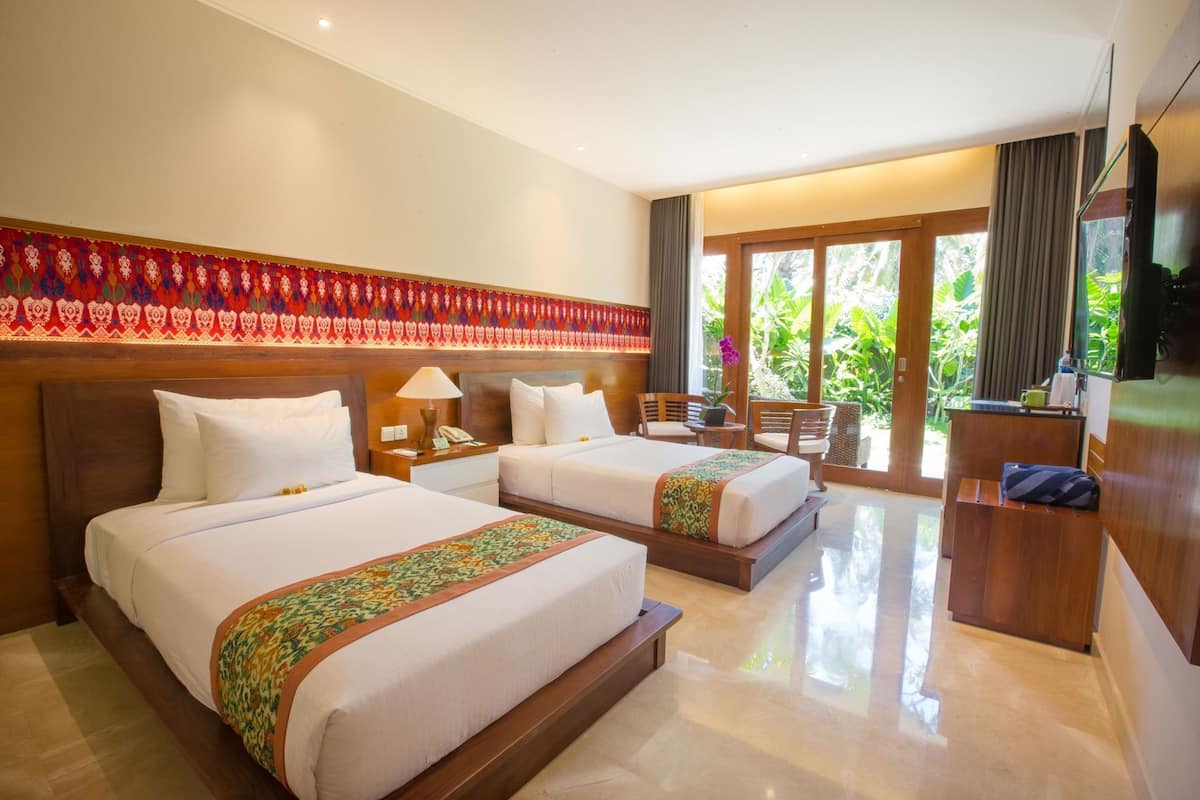 Quarto de hotel com duas camas, área de estar com televisão e vista para o verde através de grandes janelas.