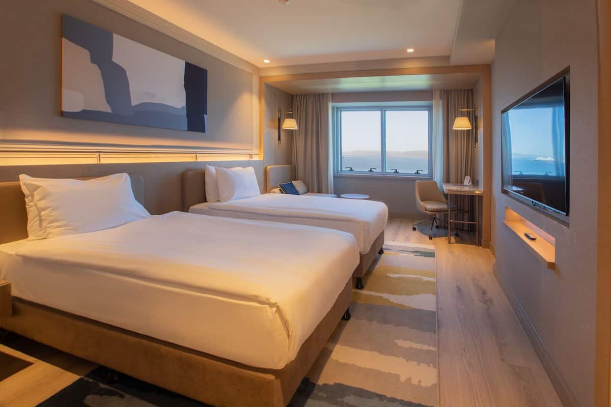 A hotel room with two beds, a desk, a chair, a TV, and a large window with a view.
