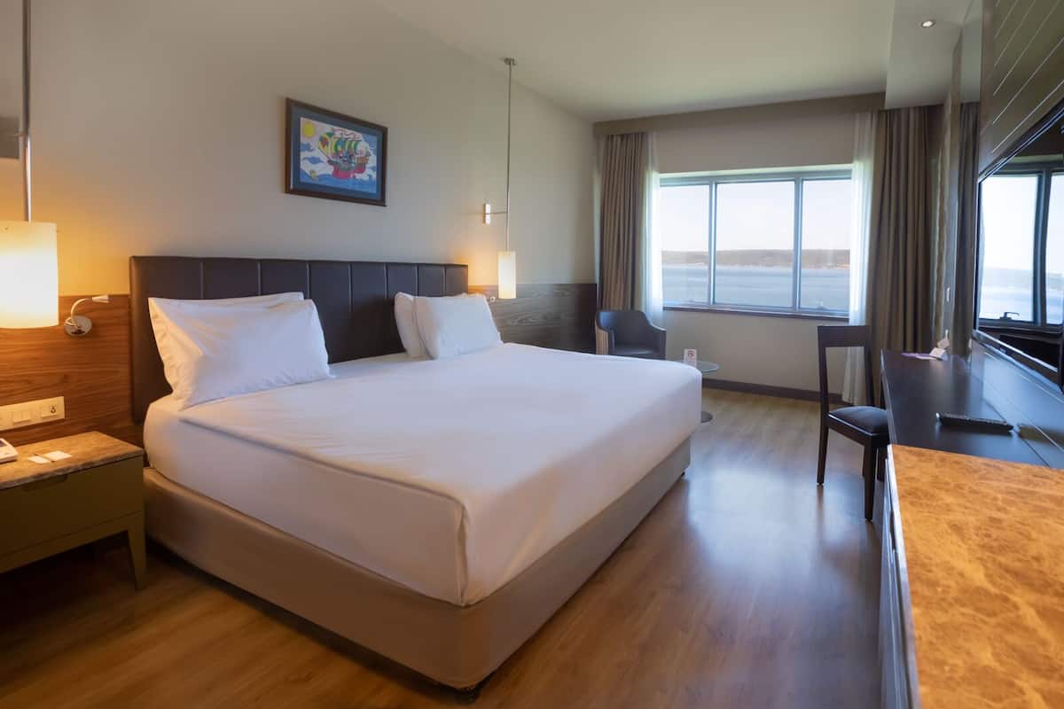 A hotel room with a large bed, a desk, a chair, a window with a view, and a painting on the wall.
