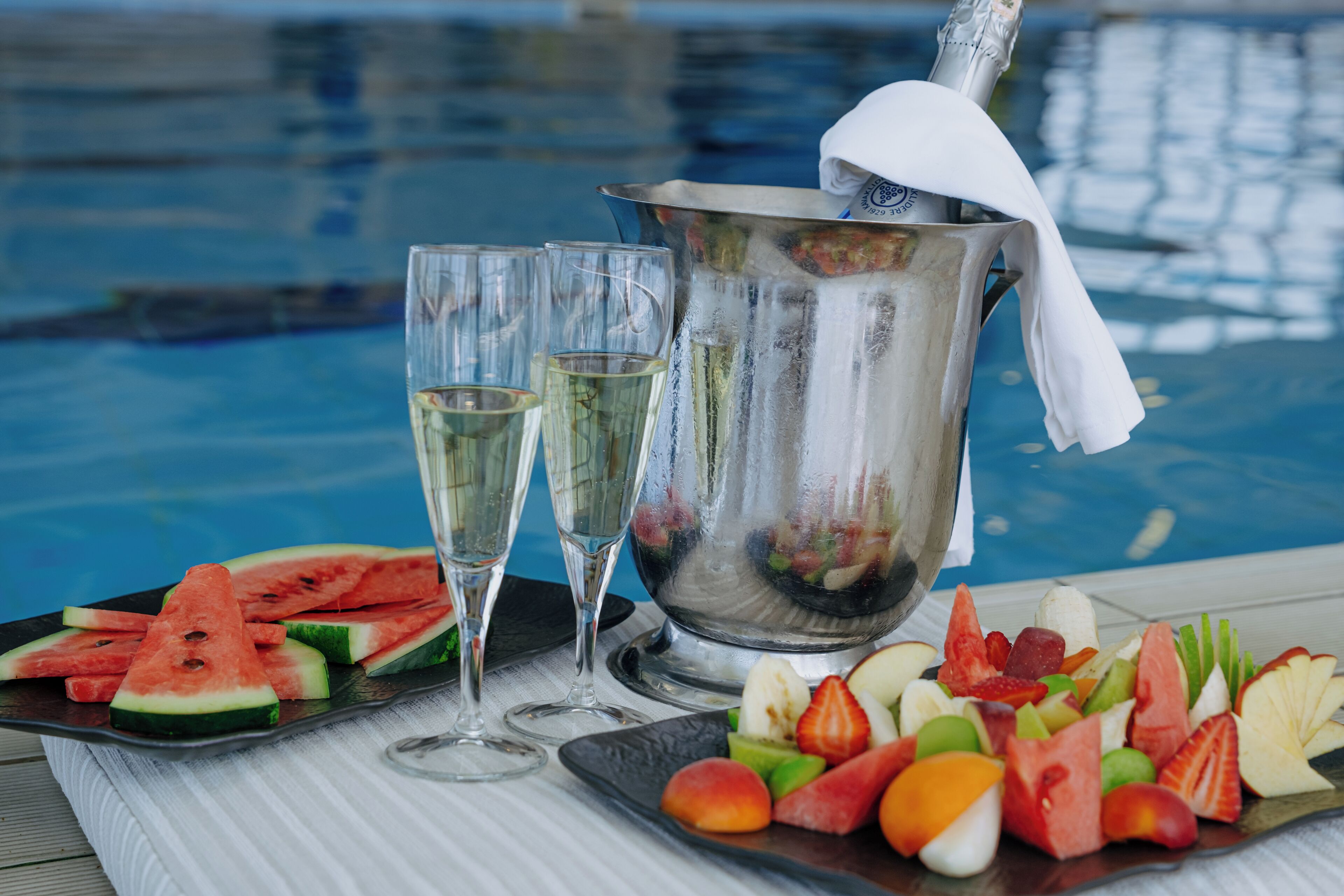 A poolside setting with a fruit platter, two glasses of champagne, and a bucket of ice.