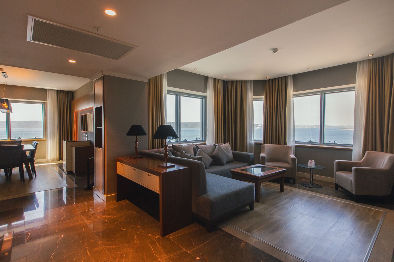 A modern hotel room with a dining area, seating arrangement, and large windows offering a view of the sea.