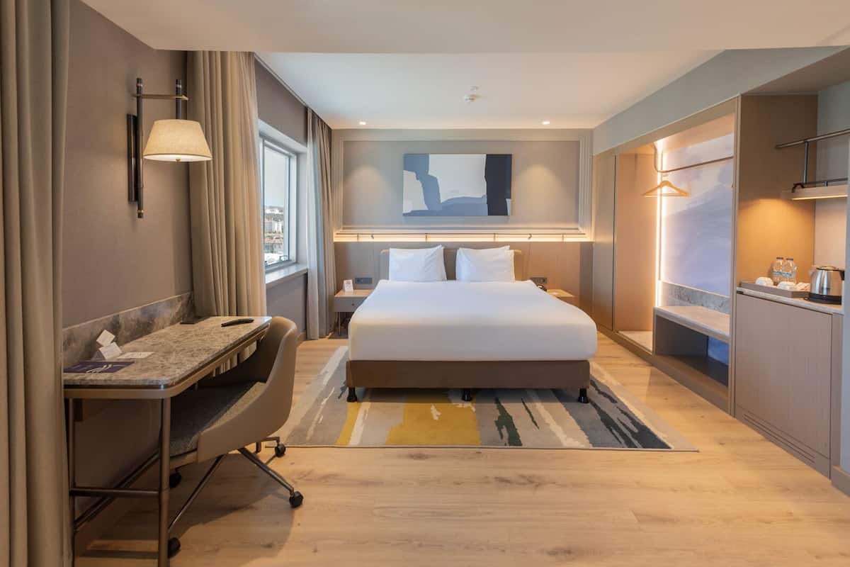 A modern hotel room with a bed, a desk, a chair, a small table, a nightstand, and a window with curtains.