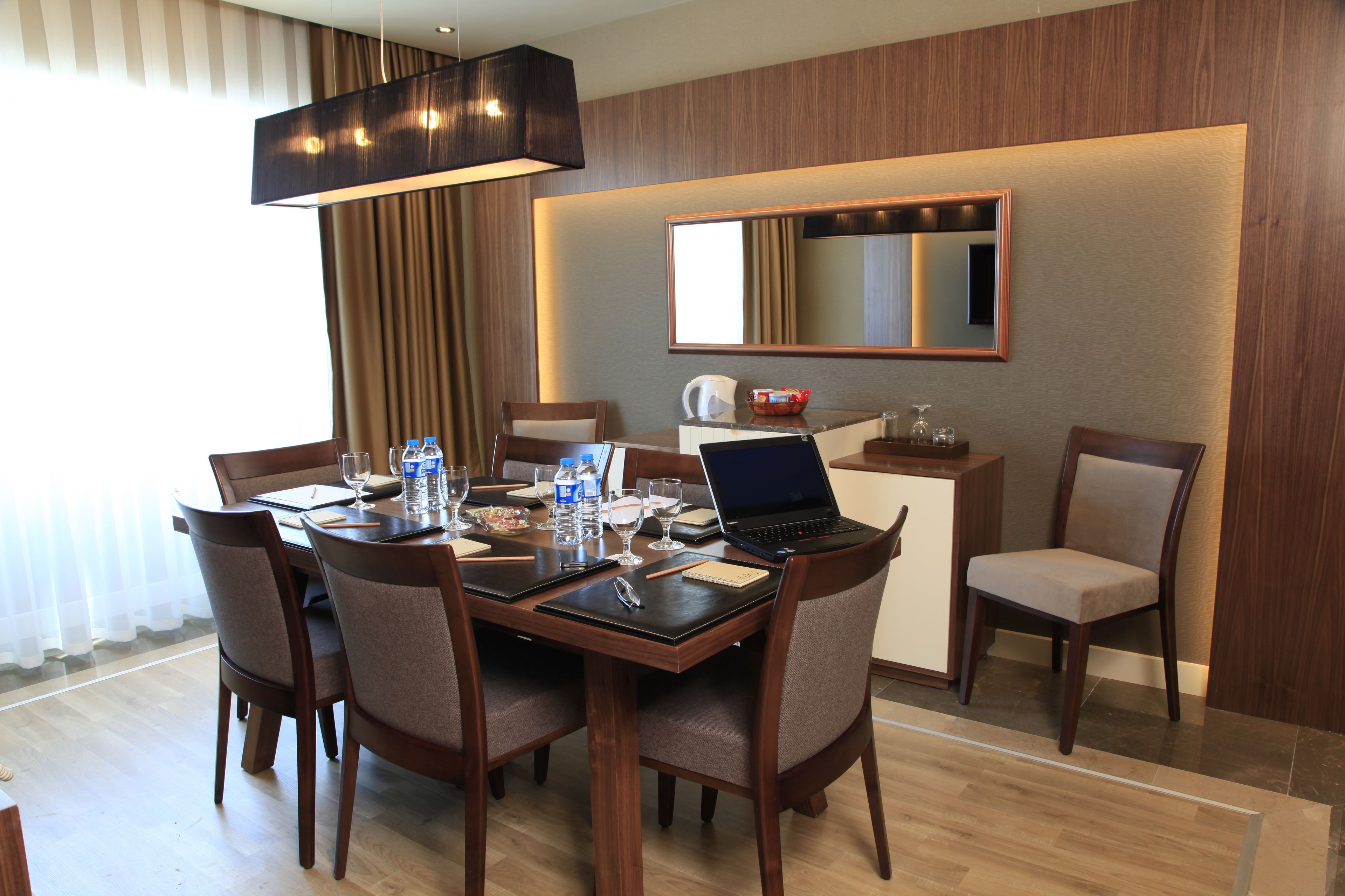 A hotel dining area with a wooden table, chairs, a laptop, and various items on the table.