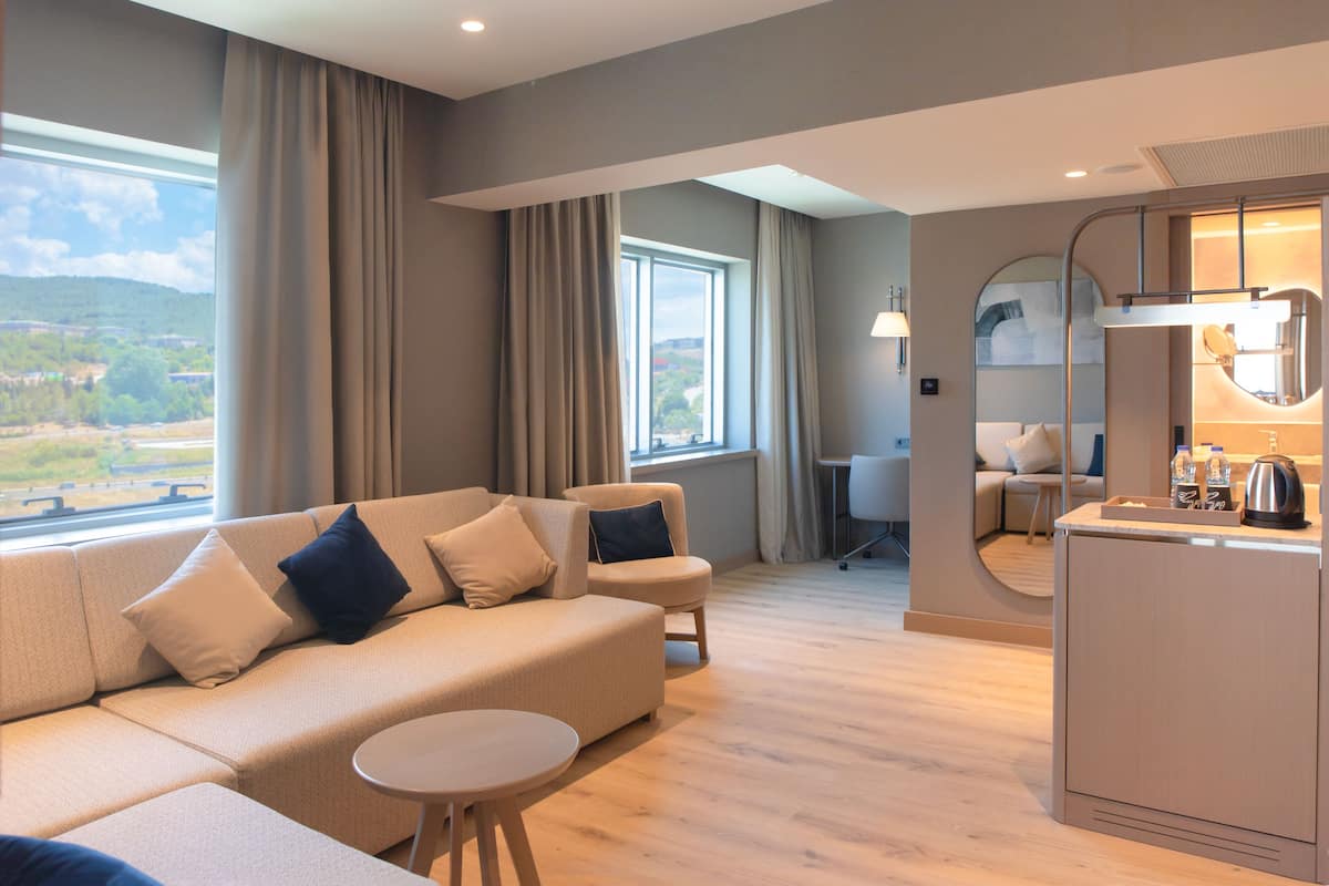 A modern hotel room with a sofa, a small round table, a desk, and a kitchenette.