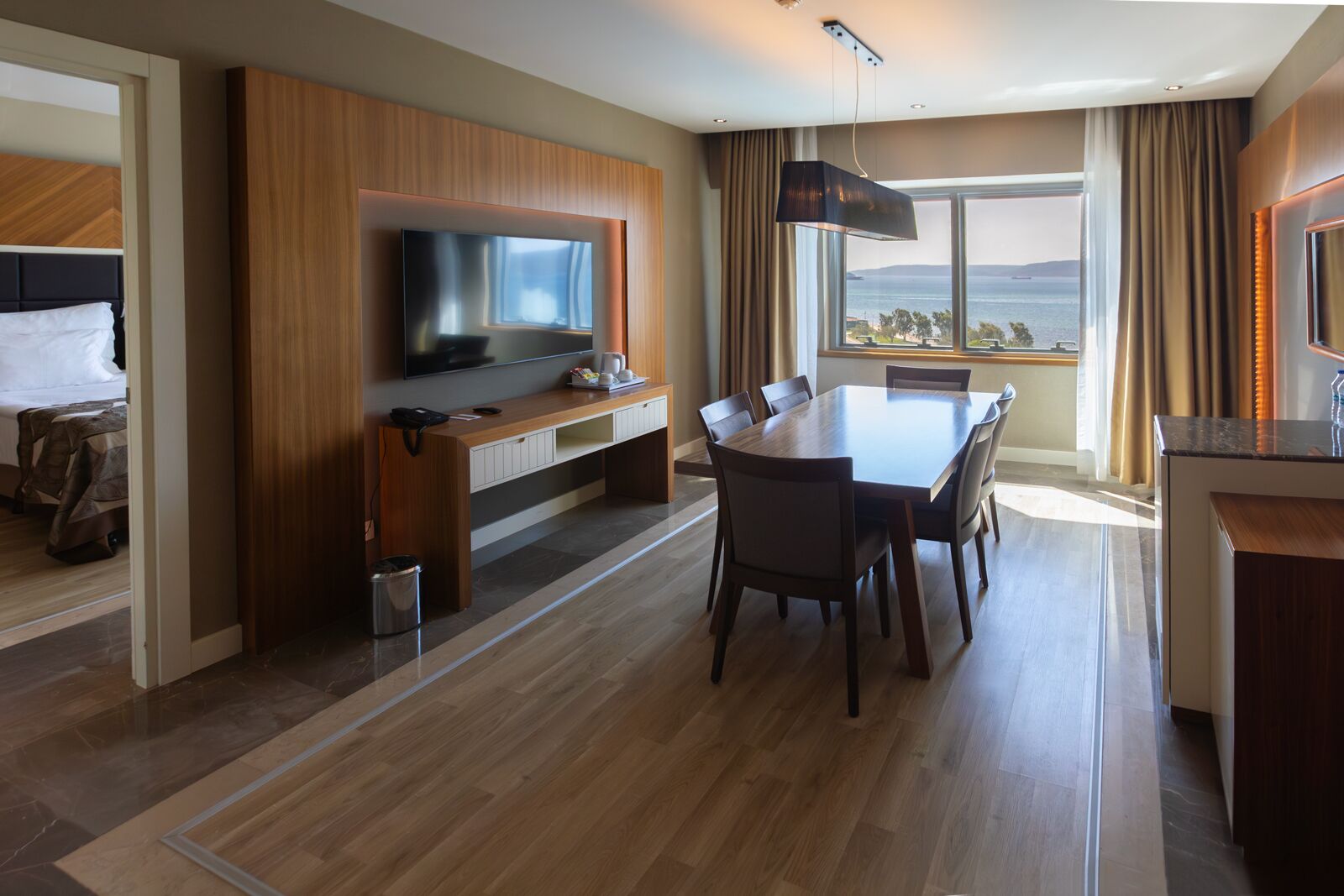 A hotel room with a wooden floor, a dining area with a table and chairs, a TV mounted on a wooden panel, and a view of the sea through large windows.