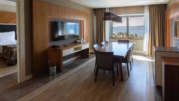 A hotel room with a wooden floor, a dining area with a table and chairs, a TV mounted on a wooden panel, and a view of the sea through large windows.