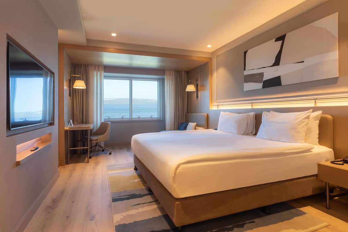 A modern hotel room with a large bed, a desk, and a view of the sea.
